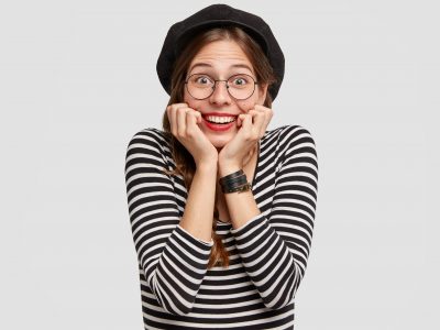 Glad Parisian female touches chin with both hands, has shining smile, cheerful facial expression, dressed in casual French style clothes, stands against white background. Positive emotions concept